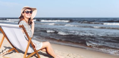 Mutlu esmer kadın güneş gözlüğü takıyor ve şapka takıyor. Okyanus sahilinde tahta bir iskemlede dinleniyor..