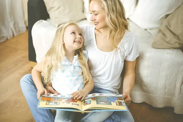 Happy family. Blonde young mother reading a book to her cute daughter while sitting at wooden floor. Motherhood concept.