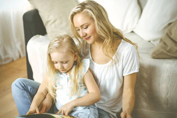 Happy family. Blonde young mother reading a book to her cute daughter while sitting at wooden floor. Motherhood concept.