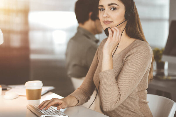Young friendly girl in headsets is talking to a firms client, while sitting at the desk in sunny office. Call center operators at work.