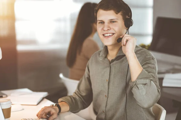 Young dark-haired guy with headsets is talking to a client, while sitting in sunny office. Call center operators at work.