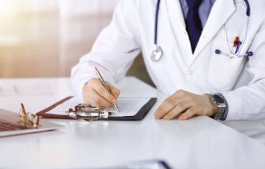 Bilinmeyen bir erkek doktor, karanlık bir klinikte tıbbi geçmişi olan bir panoyla oturuyor ve çalışıyor. Arka planda ışık parıltısı var..