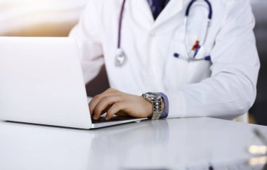 Bilinmeyen bir erkek doktor karanlık bir klinikte dizüstü bilgisayarla oturuyor ve çalışıyor. Arka planda ışık parıltısı var..