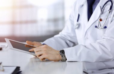 Bilinmeyen erkek doktor, tablet bilgisayarla oturuyor ve çalışıyor. Karanlık bir klinikte, arka planda ışık parıltısı, ellerin yakın çekimi..