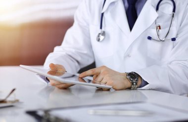 Bilinmeyen erkek doktor, tablet bilgisayarla oturuyor ve çalışıyor. Karanlık bir klinikte, arka planda ışık parıltısı, ellerin yakın çekimi..
