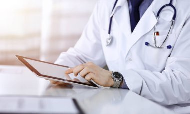 Bilinmeyen erkek doktor, tablet bilgisayarla oturuyor ve çalışıyor. Karanlık bir klinikte, arka planda ışık parıltısı, ellerin yakın çekimi..