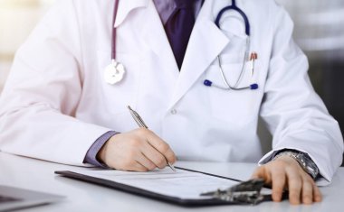 Bilinmeyen bir erkek doktor, karanlık bir klinikte tıbbi geçmişi olan bir panoyla oturuyor ve çalışıyor. Arka planda ışık parıltısı var..