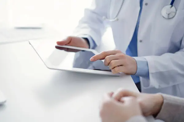 stock image Doctor and patient sitting at the table in clinic. The focus is on female physicians hands using tablet computer, close up. Medicine and healthcare concept.