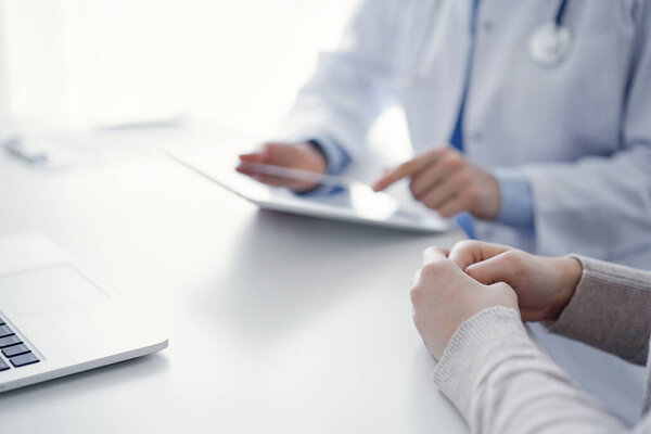 Doctor and patient sitting at the table in clinic while using tablet computer. The focus is on female patients hands, close up. Medicine concept.