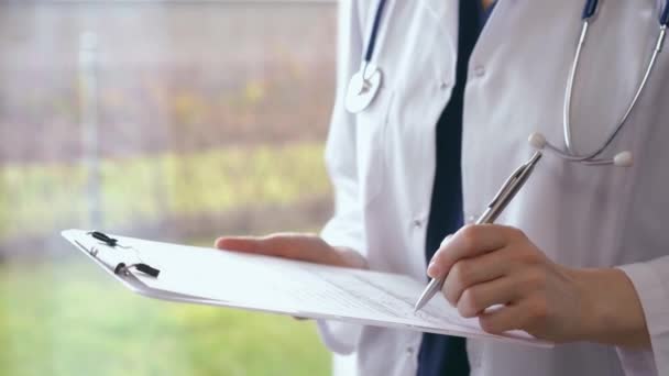 Doctor is writing something using a clipboard while standing near window in clinic, close up. Female physician or surgeon at work. Medicine concept.