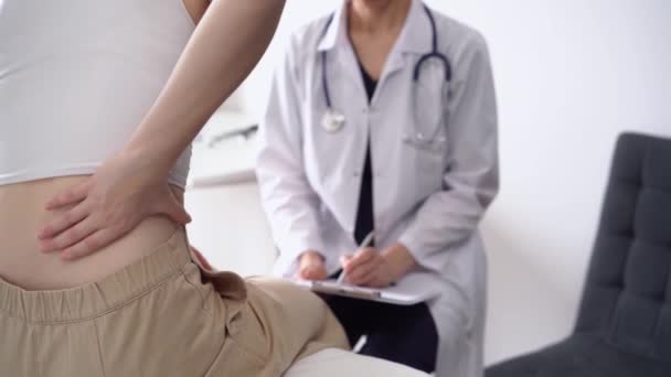 医師と患者さんは診療所にいます 女性が背中をこすっている間 メモを作るためにクリップボードを使用して白いコートの整形外科 閉じます 理学療法の概念 — ストック動画