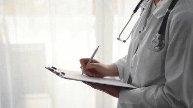 Doktor kadın pano tutarken ve klinikteki panorama penceresinin yanında dururken tıbbi kayıtları dolduruyor. Tıp konsepti.