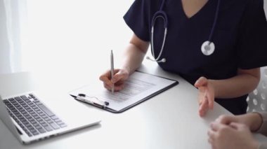 Doktor ve hasta klinikteki yetenek penceresinin yanındaki beyaz masada oturup test sonuçlarını tartışıyor. Bilinmeyen doktor tıbbi kayıt formlarını doldurmak için pano kullanıyor. Tıp konsepti.