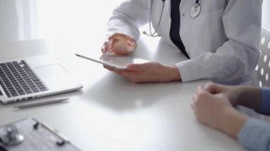 Doktor ve hasta klinikteki özel pencerenin yanındaki beyaz masada oturuyorlar. Beyaz önlük giyen kimliği belirsiz kadın doktor tıbbi kayıt formlarını doldurmak için tablet bilgisayar kullanıyor. Tıp konsepti.
