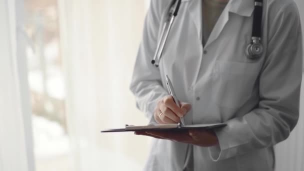 クリップボードを持ち クリニックのパノラマウィンドウの近くに立っている間に医学記録を埋める医師の女性 医学コンセプト — ストック動画