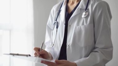 Doktor kadın, tıbbi notlar yazarken beyaz bir not panosu kullanıyor. Hastanenin panorama penceresinin yanında duran bilinmeyen bir doktor. İlaç..