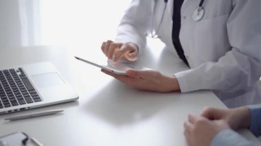 Doktor ve hasta klinikteki özel pencerenin yanındaki beyaz masada oturuyorlar. Beyaz önlük giyen kimliği belirsiz kadın doktor tıbbi kayıt formlarını doldurmak için tablet bilgisayar kullanıyor. Tıp konsepti.