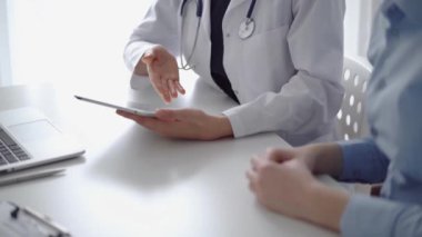 Doktor ve hasta klinikteki özel pencerenin yanındaki beyaz masada oturuyorlar. Beyaz önlük giyen kimliği belirsiz kadın doktor tıbbi kayıt formlarını doldurmak için tablet bilgisayar kullanıyor. Tıp konsepti.