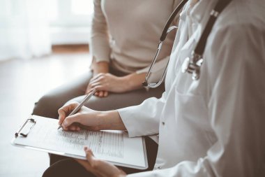 Doktor ve hasta klinikteki koltukta oturuyor. Kadın doktorların ellerinin ilaç geçmişi formunu doldurmasına odaklan, kapat. Tıp konsepti.