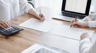 Ofisteki ahşap masada vergileri sayarken iki muhasebeci dizüstü bilgisayar ve hesap makinesi kullanıyor. İş denetimi ve finans alanında takım çalışması.