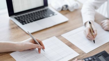 Ofisteki ahşap masada vergileri sayarken iki muhasebeci dizüstü bilgisayar ve hesap makinesi kullanıyor. İş denetimi ve finans alanında takım çalışması.