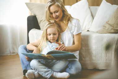 Mutlu bir aile. Sarışın genç anne ahşap zeminde otururken sevimli kızına kitap okuyor. Annelik kavramı.
