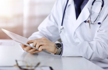 Bilinmeyen erkek doktor, tablet bilgisayarla oturuyor ve çalışıyor. Karanlık bir klinikte, arka planda ışık parıltısı, ellerin yakın çekimi..