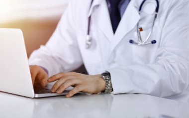 Bilinmeyen bir erkek doktor karanlık bir klinikte dizüstü bilgisayarla oturuyor ve çalışıyor. Arka planda ışık parıltısı var..