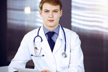 Sarışın erkek doktor iş yerindeki güneşli klinikte tablet bilgisayarla oturup çalışıyor. Yakın plan. Tıp konsepti.