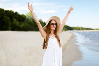 Okyanus sahilinde mutlu, mutlu, mutlu bir kadın şapkası, güneş gözlüğü ve fırfırlı elleriyle duruyor. Beyaz yaz elbisesi içinde doğanın tadını çıkaran çok kültürlü bir kadın modelin portresi.