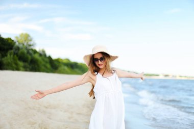 Okyanus sahilinde mutlu, mutlu, mutlu bir kadın şapkası, güneş gözlüğü ve açık elleriyle duruyor. Beyaz yaz elbisesi içinde çok kültürlü bir kadın modelin portresi seyahat sırasında doğanın tadını çıkarıyor.