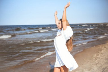 Mutlu gülümseyen güzel bir kadın okyanus sahilinde beyaz bir yaz elbisesi içinde el kaldırıyor..