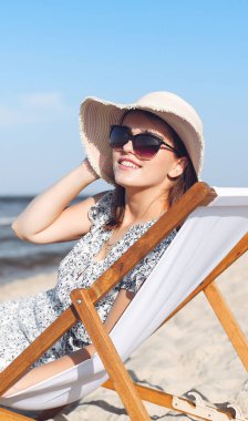 Mutlu esmer kadın güneş gözlüğü takıyor ve şapka takıyor. Okyanus sahilinde tahta bir iskemlede dinleniyor..