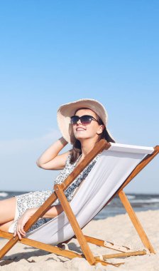 Mutlu esmer kadın güneş gözlüğü takıyor ve şapka takıyor. Okyanus sahilinde tahta bir iskemlede dinleniyor..