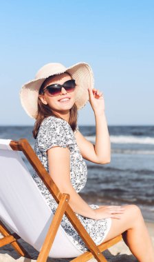 Okyanus sahilinde tahta bir iskemlede dinlenirken güneş gözlüğü takan mutlu esmer kadın..