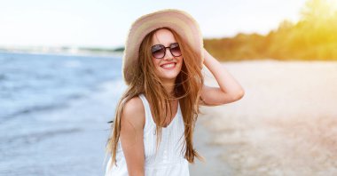 Okyanus sahilinde mutluluk içinde gülümseyen mutlu kadın şapka ve güneş gözlükleriyle poz veriyor. Beyaz yaz elbisesi içinde doğanın tadını çıkaran bir kadın modelin portresi.