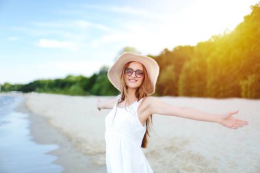 Okyanus sahilinde mutlu, mutlu, mutlu bir kadın şapkası, güneş gözlüğü ve açık elleriyle duruyor. Beyaz yaz elbisesi içinde, doğanın tadını çıkaran çok kültürlü bir kadın modelin portresi..