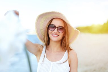 Okyanus sahilinde mutluluk içinde gülümseyen mutlu bir kadın şapka, güneş gözlüğü ve beyaz çiçeklerle duruyor. Beyaz yaz elbisesi içinde, doğanın tadını çıkaran çok kültürlü bir kadın modelin portresi..