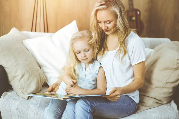 Happy family. Blonde young mother reading a book to her cute daughter while sitting at wooden floor in sunny room. Motherhood concept.