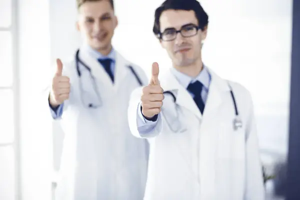 2人の男性医師がチームとして立ち 診療所の患者にとって最高のサービスの象徴として親指を立てています ヘルスケア — ストック写真