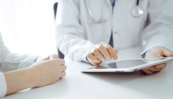 stock image Doctor and patient sitting near each other at the desk in clinic. The focus is on female physicians hands pointing into tablet computer touchpad, close up. Medicine concept.