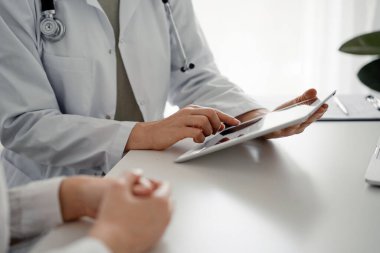 Doktor ve hasta klinikteki masada oturuyorlar. Tablet bilgisayar kullanan kadın doktorların ellerine odaklan, kapat. Mükemmel tıbbi hizmet ve tıp konsepti.