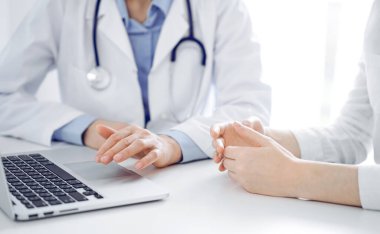Doktor ve hasta klinikteki masada yan yana oturuyorlar. Diz üstü bilgisayarı kullanan kadın doktorların ellerine odaklan, kapat. Tıp konsepti.