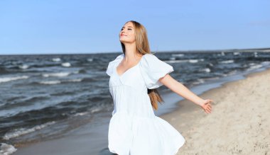 Mutlu gülümseyen güzel kadın okyanus sahilinde beyaz bir yaz elbisesi içinde, kolları açık..