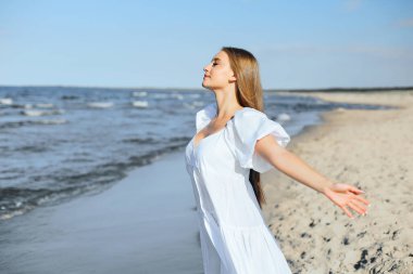Mutlu gülümseyen güzel kadın okyanus sahilinde beyaz bir yaz elbisesi içinde, kolları açık..