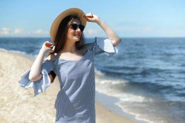 Okyanus sahilinde gülümseyen, moda şapkası ve güneş gözlüğü takan mutlu, genç esmer kadın. Keyifli tatil konsepti.