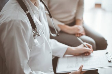 Doktor ve hasta klinikteki koltukta oturuyor. Kadın doktorların ellerinin ilaç geçmişi formunu doldurmasına odaklan, kapat. Tıp konsepti.
