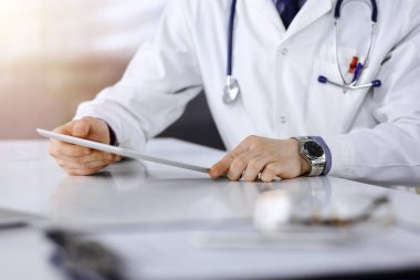 Bilinmeyen erkek doktor, tablet bilgisayarla oturuyor ve çalışıyor. Karanlık bir klinikte, arka planda ışık parıltısı, ellerin yakın çekimi..