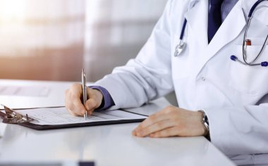 Bilinmeyen bir erkek doktor, karanlık bir klinikte tıbbi geçmişi olan bir panoyla oturuyor ve çalışıyor. Arka planda ışık parıltısı var..