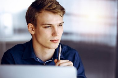 Genç sarışın işadamı, çalışma yerinde, karanlık bir ofiste bilgisayarla strateji düşünüyor. Arka planda ışık parıltısı var..